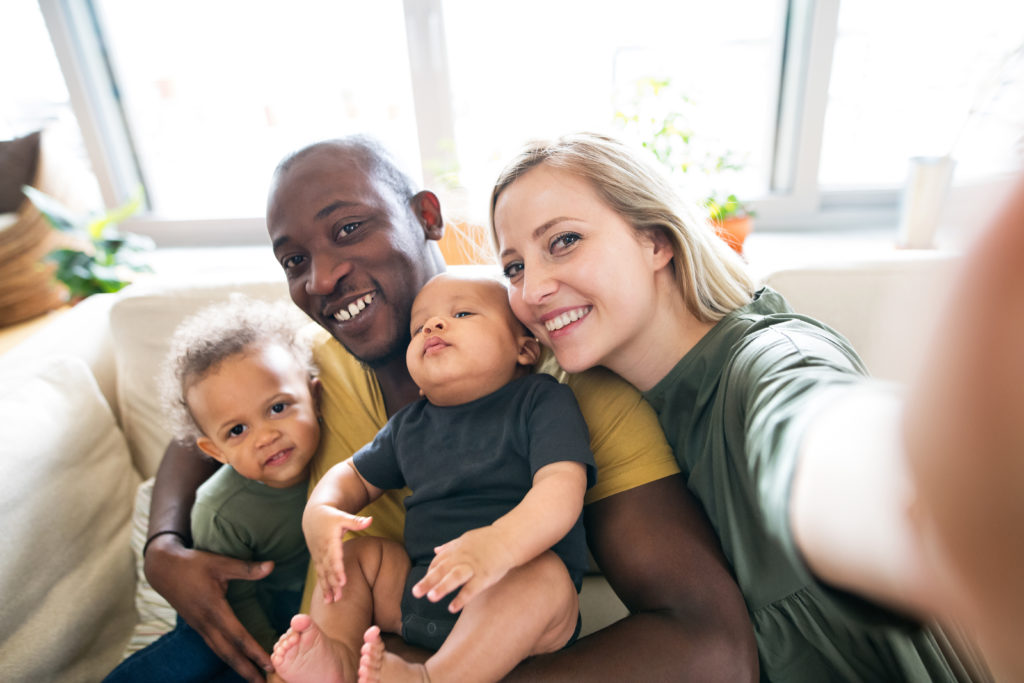 Beautiful young interracial family at home holding their cute son and daughter in the arms, taking selfie.