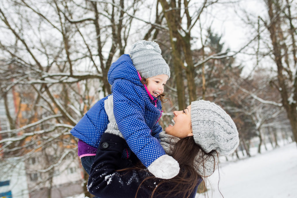 Beautiful young mother with her cute little daughter playing outside in winter nature. Mom lifting girl up.